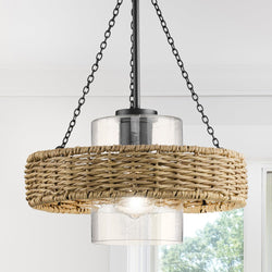 The UHP4780 Casual Indoor Pendant from the Urban Ambiance Bexley Collection showcases a weather-resistant rattan shade with a clear glass cylinder protecting the bulb, all hanging from a matte black chain. Through the window in the background, lush green scenery can be seen.