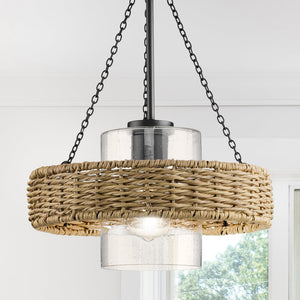 The UHP4780 Casual Indoor Pendant from the Urban Ambiance Bexley Collection showcases a weather-resistant rattan shade with a clear glass cylinder protecting the bulb, all hanging from a matte black chain. Through the window in the background, lush green scenery can be seen.